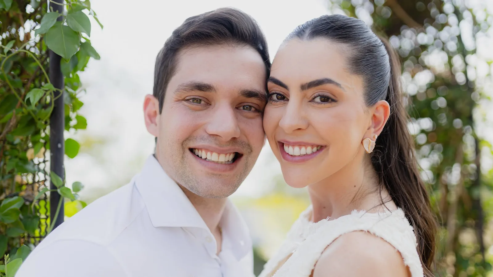Leandro e Laís rosto com rosto, sorrindo, com fundo claro e folhagem desfocada